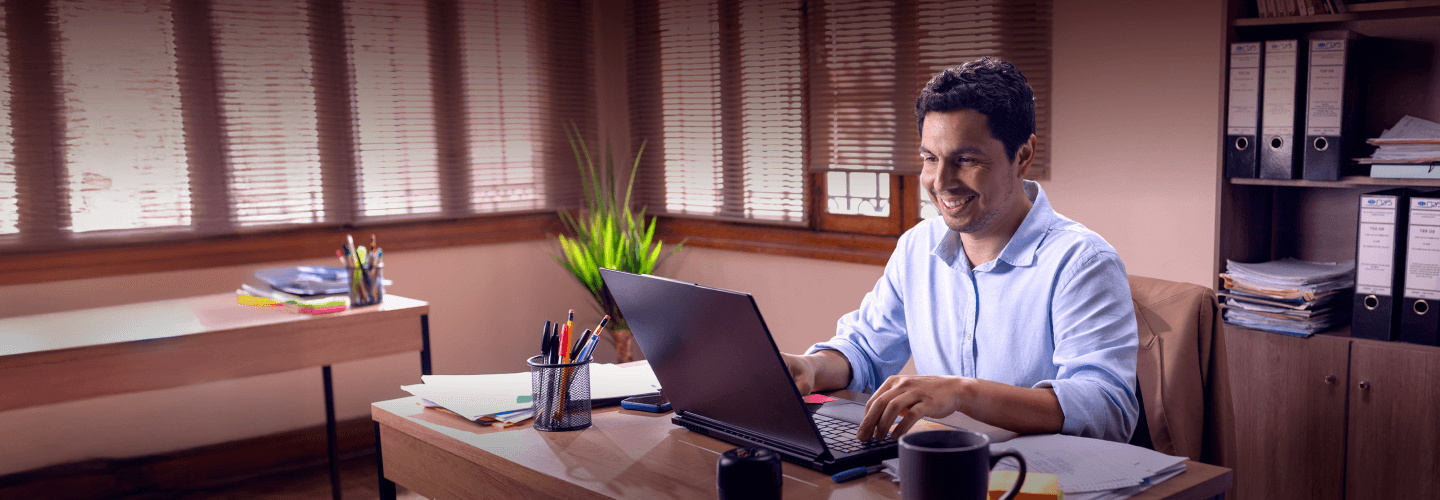 Hombre sonriendo mientras realiza operaciones desde su laptop y descubre que sus ahorros crecen con la Cuenta WOW de Compartamos Banco, con 4.60% TREA en soles.